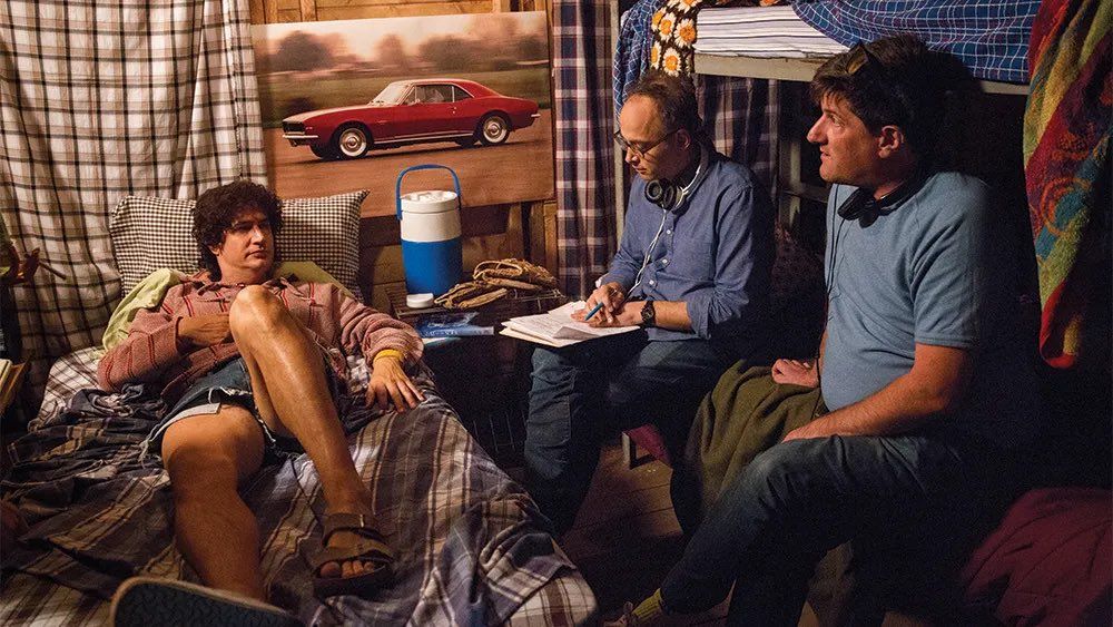 A behind the scenes photo from the Netflix series Wet Hot American Summer: First Day of Camp: an actor lies on a bed and the director and writer discuss a scene with him