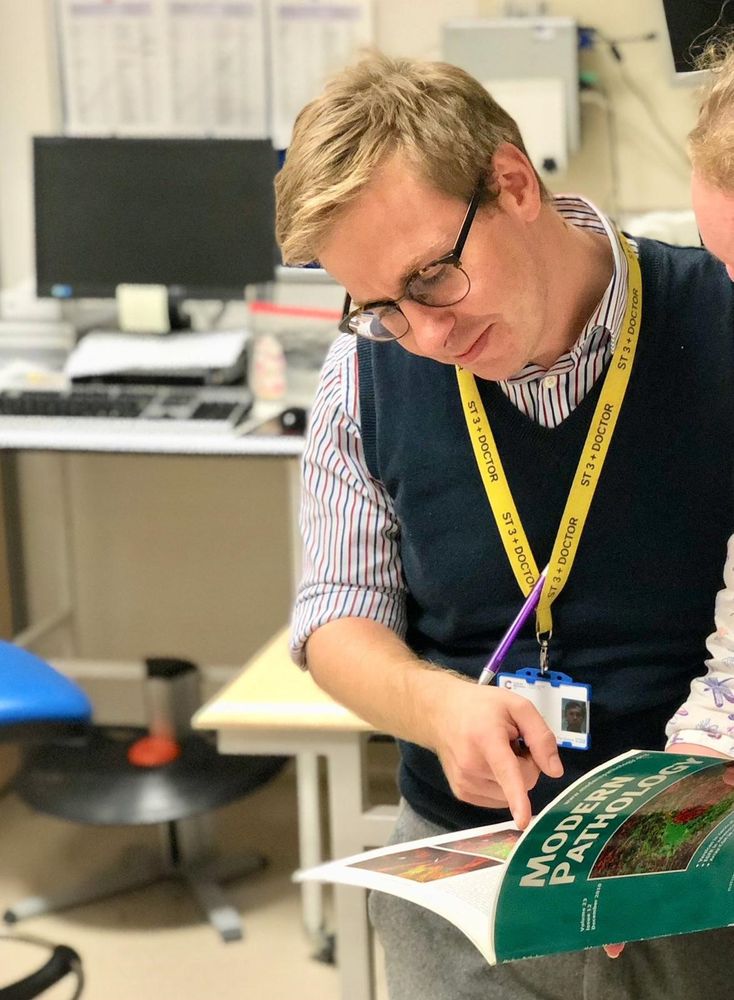 Dr Neil Ryan, stood in a lab/office space, pointing at a book on modern pathology that a colleague is holding