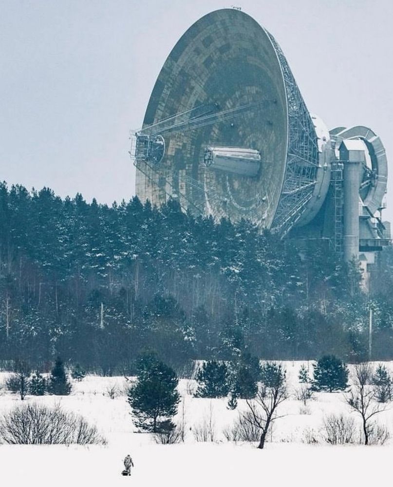 Photo of the telescope in winter. A massive, hulking, brutal radio dish rises from behind a thick row of coniferous trees. A person, minuscule by comparison, is in the foreground.