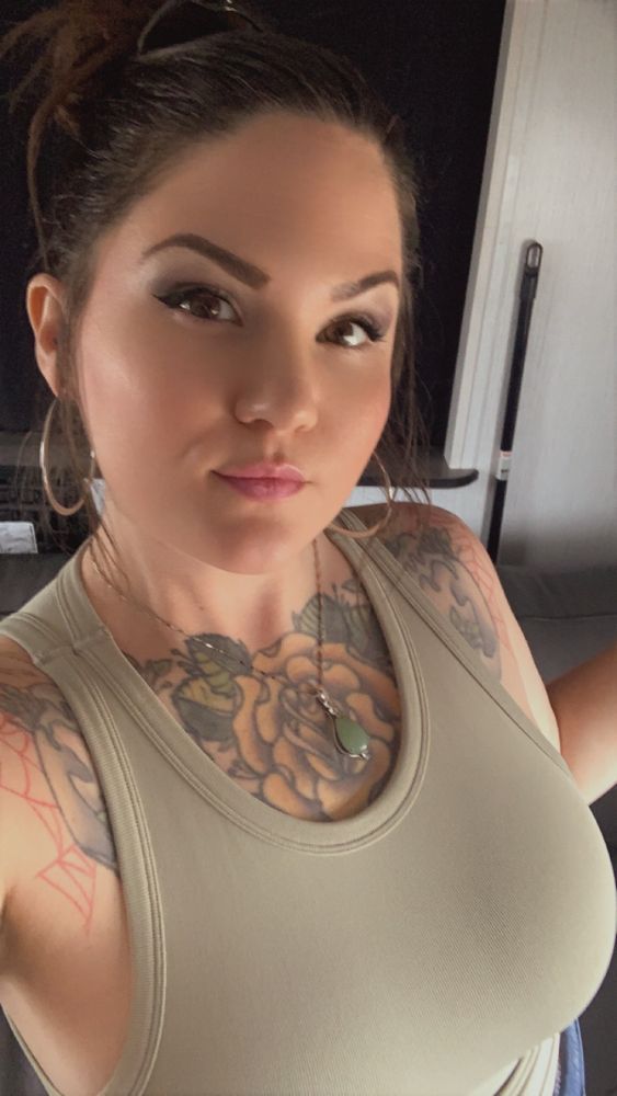 Tattooed brunette taking a selfie, wearing a light olive tank top