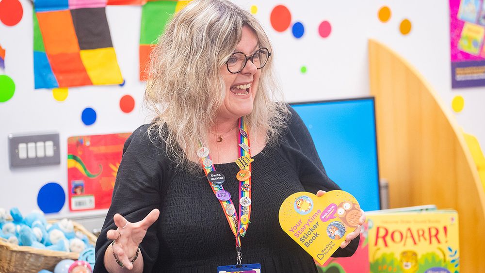 A librarian smiling while leading a BookTrust Storytime session, holding a BookTrust Storytime sticker book
