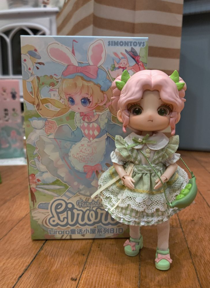 Photo of a Liroro Fairytale House doll, about six inches tall with pink hair and a green dress. She looks grumpy and her purse is a pea pod.