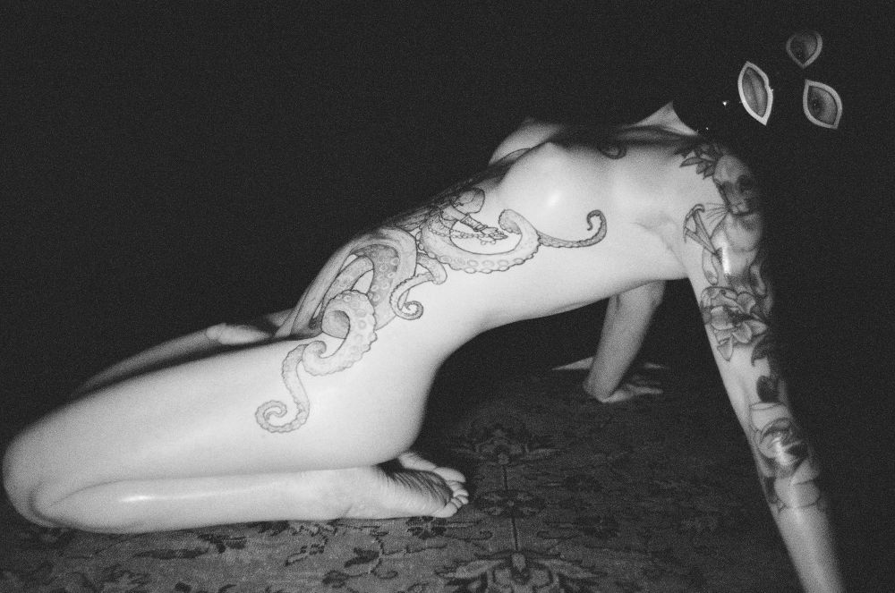 B&W 35mm film image of a tattooed trans woman nude except for a black latex hood in a dark room sitting on her knees bending back with her tits up resting on her hands behind her.