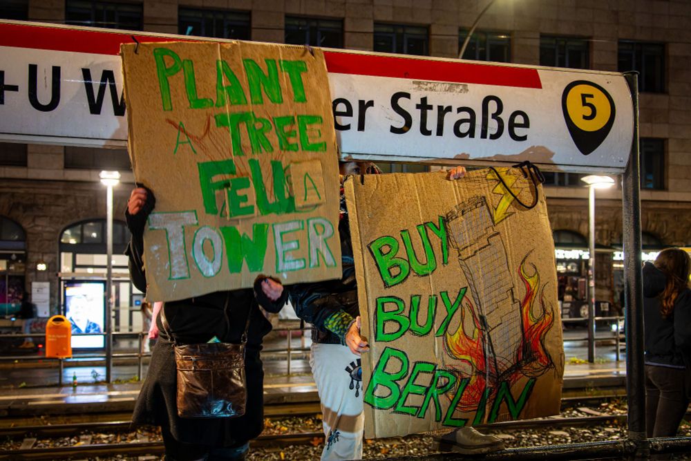 An der Tramhaltestelle halten 2 Menschen selbstgemalte Schilder hoch.
"Plant a Tree, fell a Tower" und "Buy Buy Berlin" mit einem gemalten Turm, der brennt.