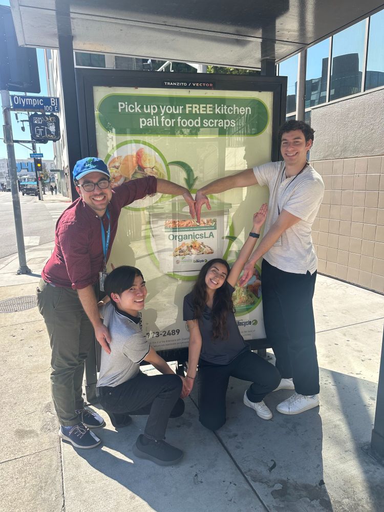 The photo shows 4 of LASAN's staff standing in front of LASAN's organics ad on a City bus shelter. They're forming a heart together around the organics pail on the sign with their arms.