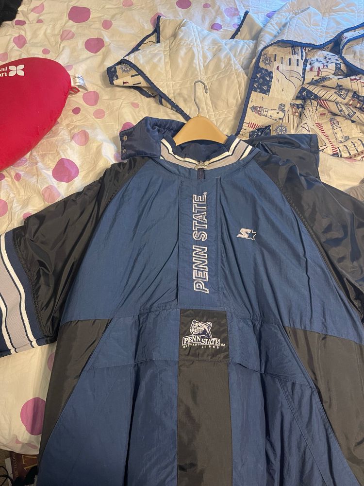 A picture of the front of a Penn State vintage Starter jacket from the late 1990s/early 2000s. It’s navy blue, with black shoulders and sides, and grey and white striping on the neck and elbows, with “PENN STATE” on the zipper cover and the turn of the millennium PSU logo on the front. 