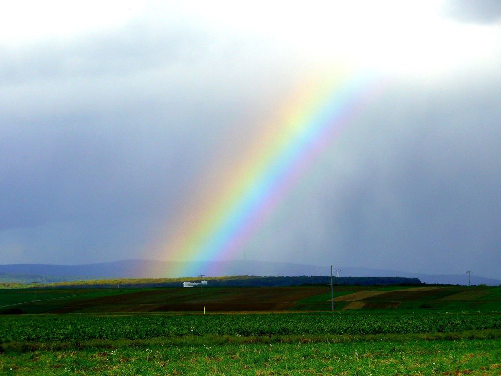 Felder und Berge in der Ferne, in der Mitte ein Regenbogen von oben nach unten 