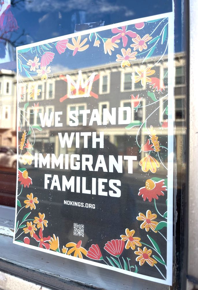 a store window with a small sign that says "we stand with immigrant families, nokings.org" — design is a black background with bright red and yellow flowers and a crown crossed out