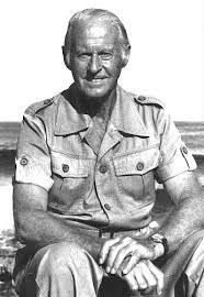 A picture of and older Thor Heyerdahl (looking a little like Joe Biden), wearing a shirt with far too many buttons. His hands are crossed on his legs. He's outside, squinting a little in the sunlight.