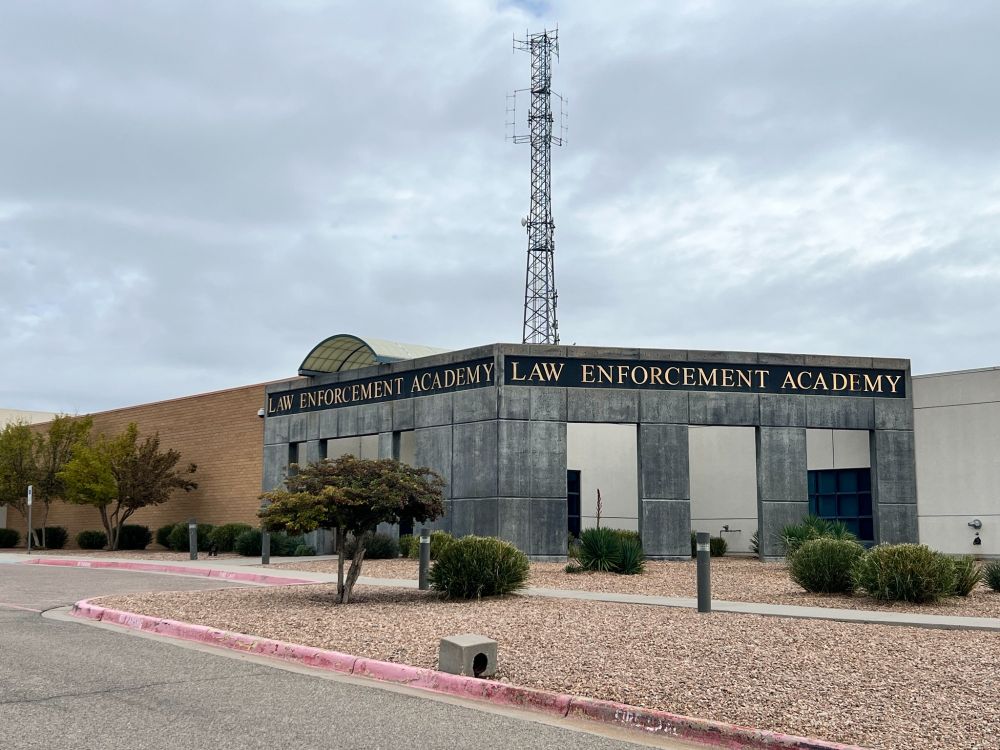 Gray block cement building with brick side with label "Law Enforcement Academy, with gravel landscaping with green bushes and trees. Sky is gray with clouds.