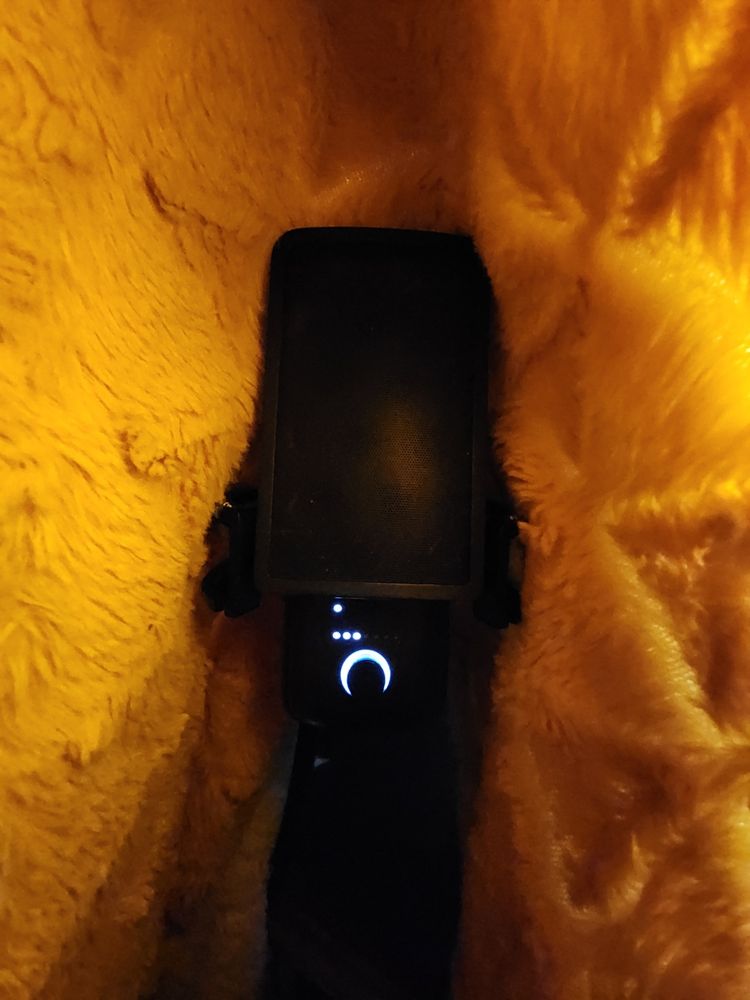 A picture of a Wave:3 microphone, covered in a golden blanket, acting as a quick and cheap sound insulation for recording vocal lines for narration purposes. A bright light shines through it to illuminate the scene, as it's normally very dark.