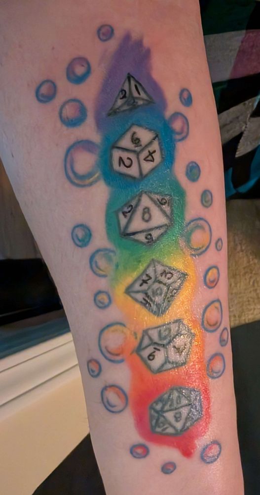 Another photo of August's forearm showing the same tattoo touched up. The "flame" design has been smoothed out, the dice have all their numbers on them now, and there are translucent bubbles surrounding the rainbow color block.