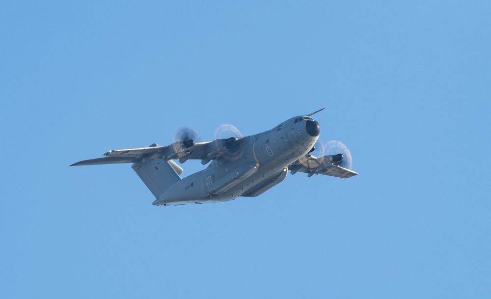 A grey military transport plane with 4 turboprop engines flies over the photographer against some lovely blue sky