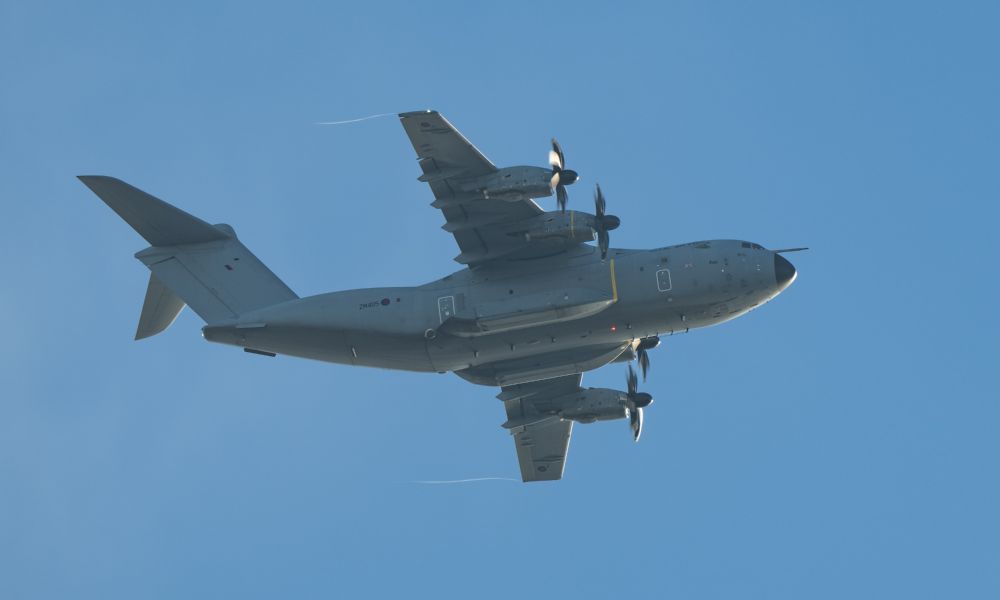 A grey military transport plane with 4 turboprop engines flies over the photographer against some lovely blue sky
