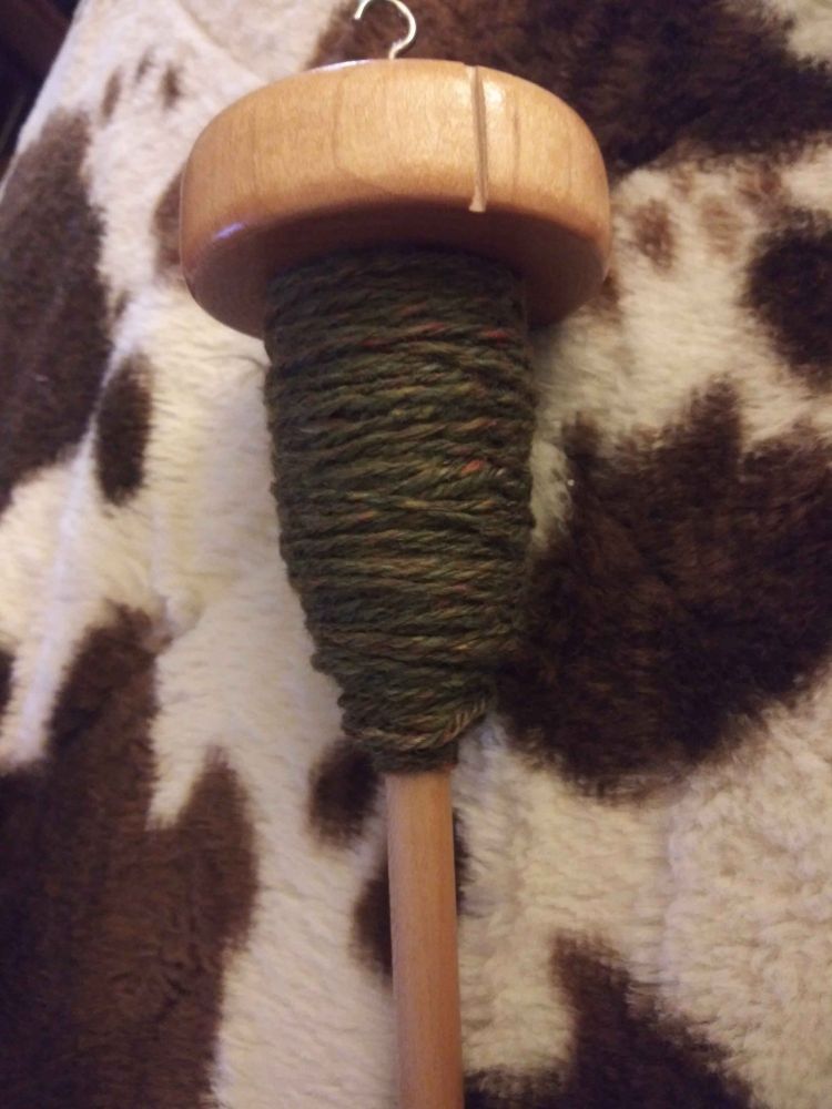 A drop spindle lays on a cow print blanket, and theres a 3 ply yarn wrapped around it. The yarn is a dark green/olive drab color with yellow, red, and blue flecks of color throughout it