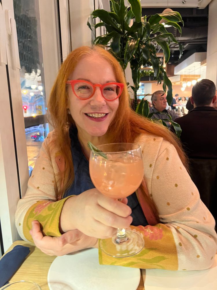 At a restaurant, a red headed, long haired white woman dressed in pink holds a big pink cocktail and smiles widely
