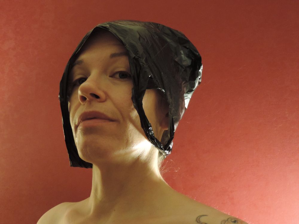 Against a painterly red-orange background, a middle-aged white woman looks imperiously down at the camera. We only see her from the shoulders up, and she seems to be naked. She is wearing a black plastic bag on her head. We see a glimpse of an octopus tattoo on her left shoulder. 