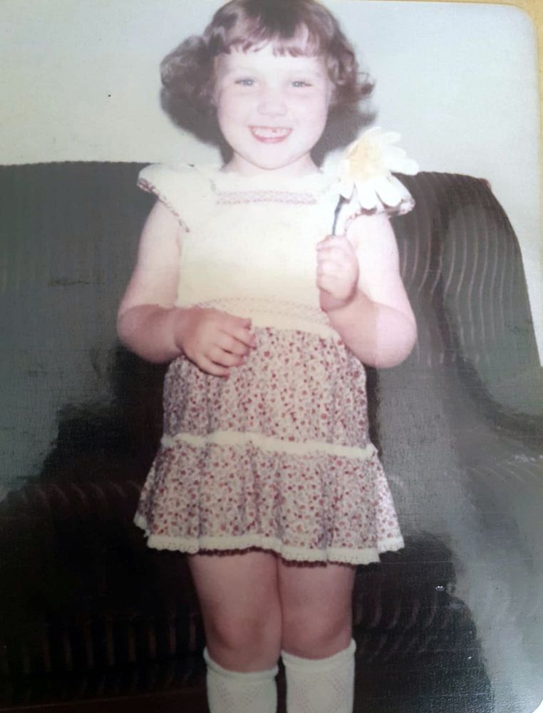 An old photo of a young girl with blonde curls wearing a 70s dress and holding a flower