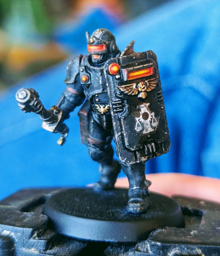 Adeptus Arbites for Warhammer 40k: Kill Team. He has black, battered armour, with a glowing red visor on his helmet and on his suppression shield. He's also armed with a shock baton. 