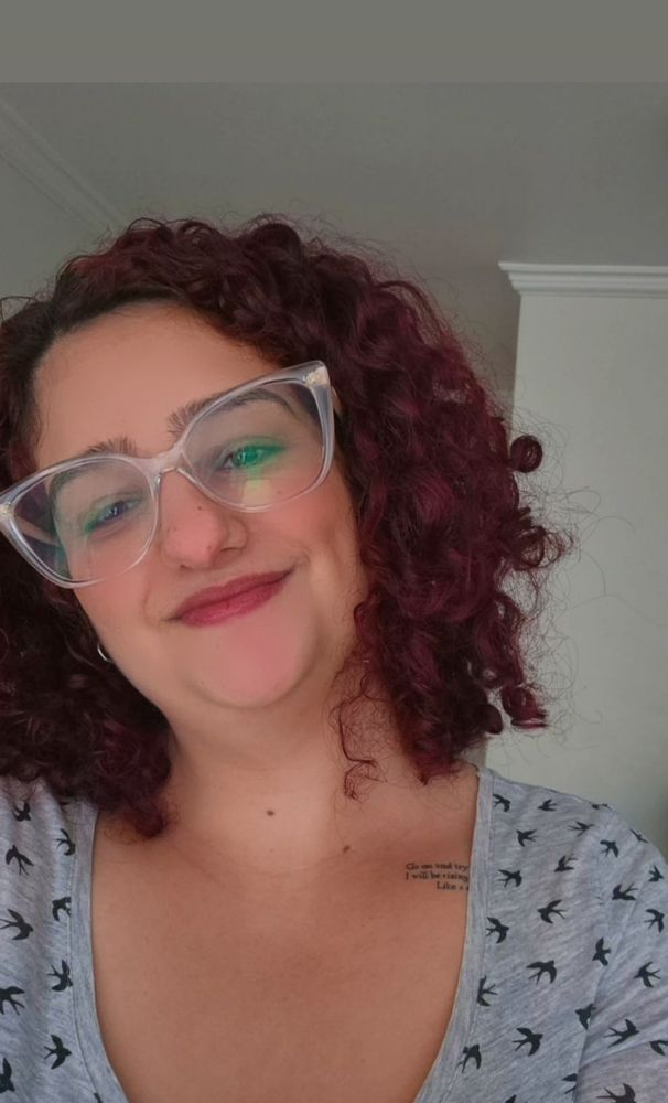 Moça de óculos, cabelo cacheado vermelho e usando blusa cinza enquanto sorri