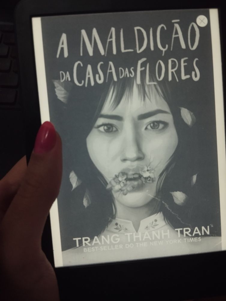 capa do livro a maldição da casa das flores: moça do Vietnã com cabelos e olhos escuros com flores ao redor dos lábios e um olhar assustado 