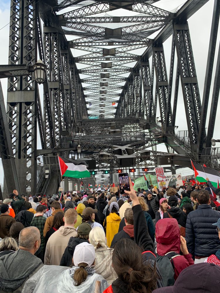 A dense crowd of protestors walking across the Sydney Harbour Bridge, waving Palestinian flags.