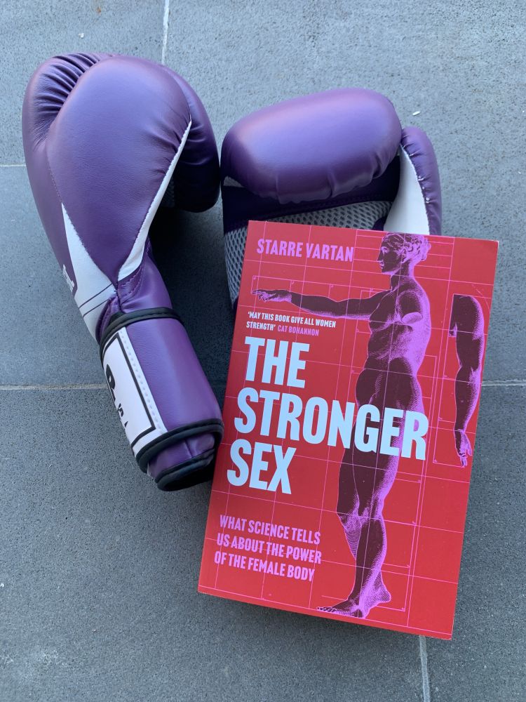 A book with a red and pink cover rests against two purple boxing gloves. The book is titled 'The Stronger Sex - What Science Tells Us About The Power Of The Female Body', by Starre Vartan.