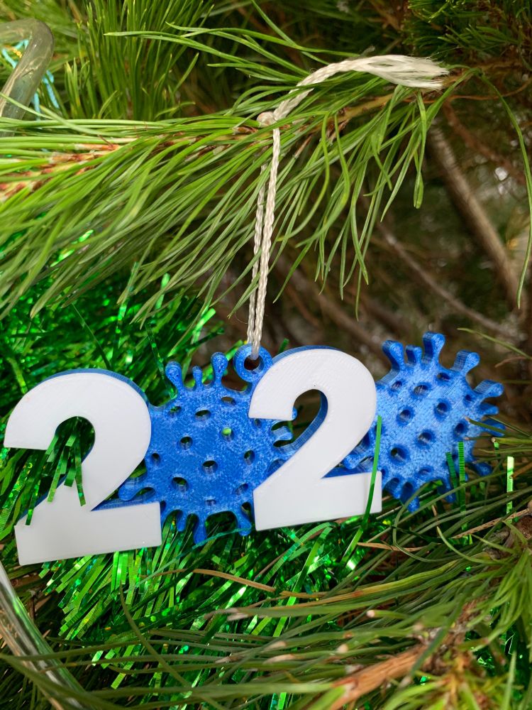 A Christmas decoration hangs in the branches of a green Christmas tree. It is the numbers 2020, with the '0's both being in the shape of blue spiky round shapes representing the SARS-CoV-2 virus.