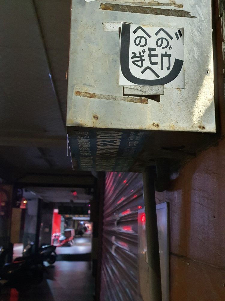 A silly little face made of graphic textual elements on a sticker that was pasted on an old utility box; below and to the side an empty sidewalk glows red, blue, and purple  reflecting off closed metal shutters.