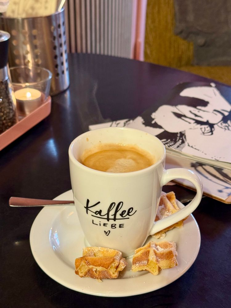 Kaffee in einer Tasse mit der Aufschrift „Kaffeeliebe“, dazu drei kleine Stückchen Waffel