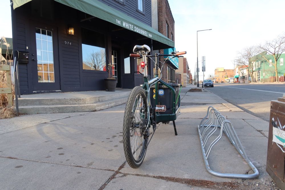 a bike stand sits on the ground next to a Bullitt cargo bike, with a bar named the White Squirrel in the background