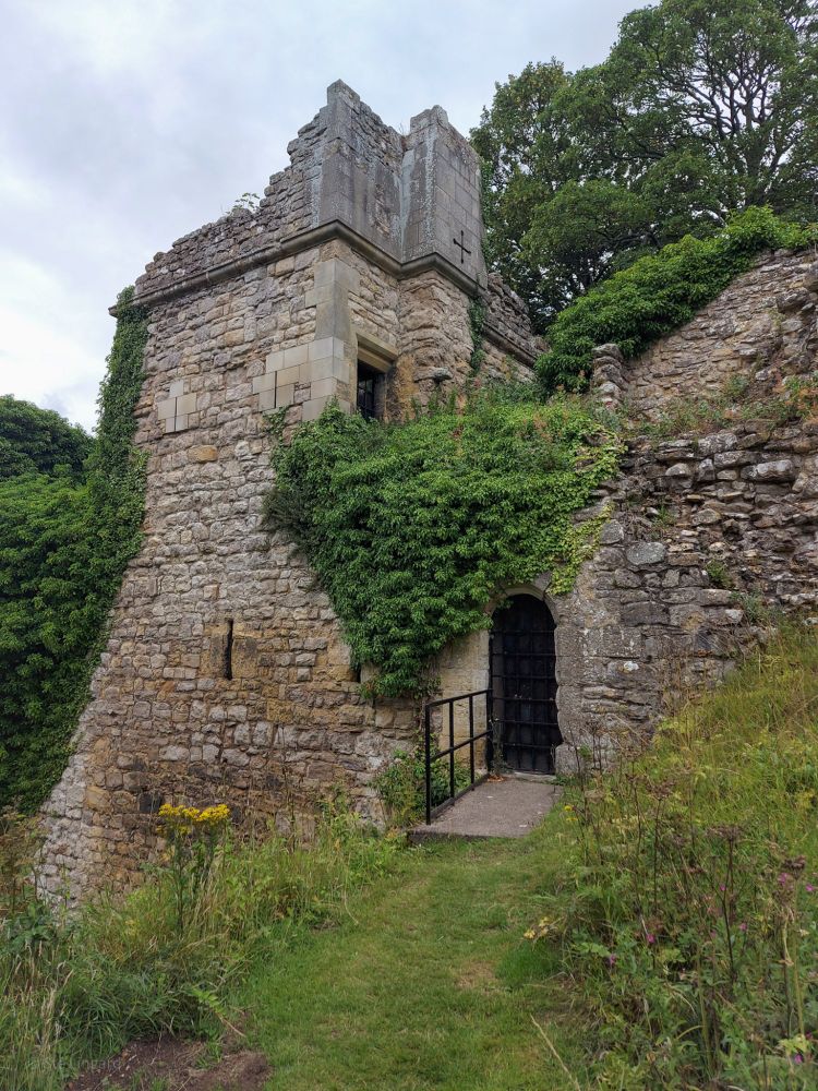 A square tower, made of stone, partly ruined and partly overgrown. 