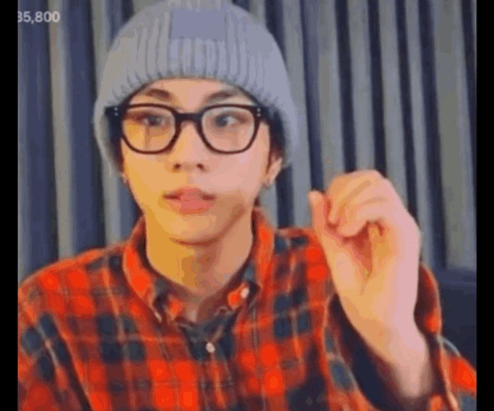 a young man wearing glasses , a plaid shirt and a beanie is making a funny face .