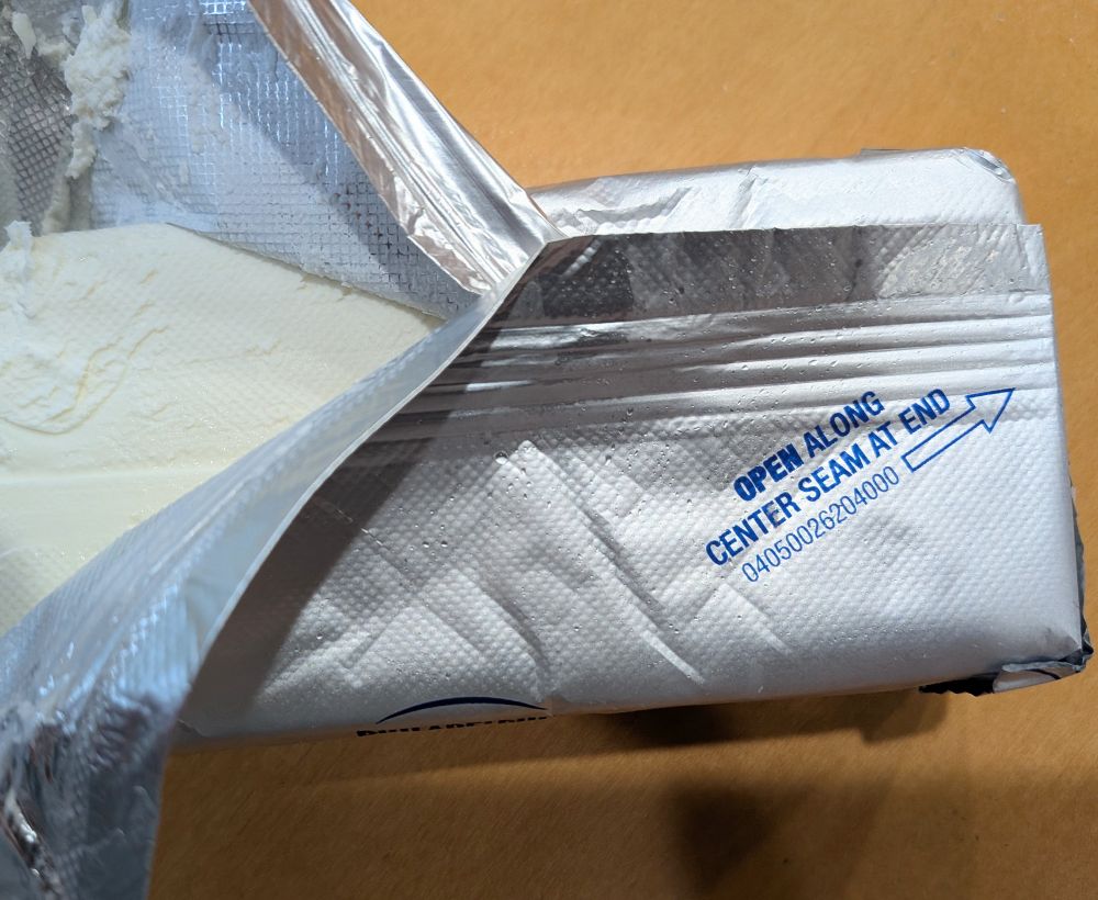 A block of Philadelphia cream cheese in the foil packaging. The instructions on the foil are "open along center seam at end" with an arrow pointing to one end, but the package is open at the opposite end the arrow is pointing.