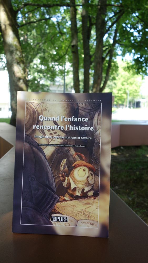 Photographie de l'ouvrage "Quand l'enfance rencontre l'histoire", posé à la verticale sur un support en forme de branche d'arbre (banc designé). C'est en extérieur, au fond, on aperçoit des arbres au feuillage flou. La couverture est dans les tons marron et jaune, et le banc est lui aussi marron.