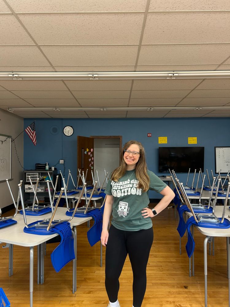 Teacher in her classroom with a shirt that says “Teaching math, expecting an addition” with an adorable baby calculator 