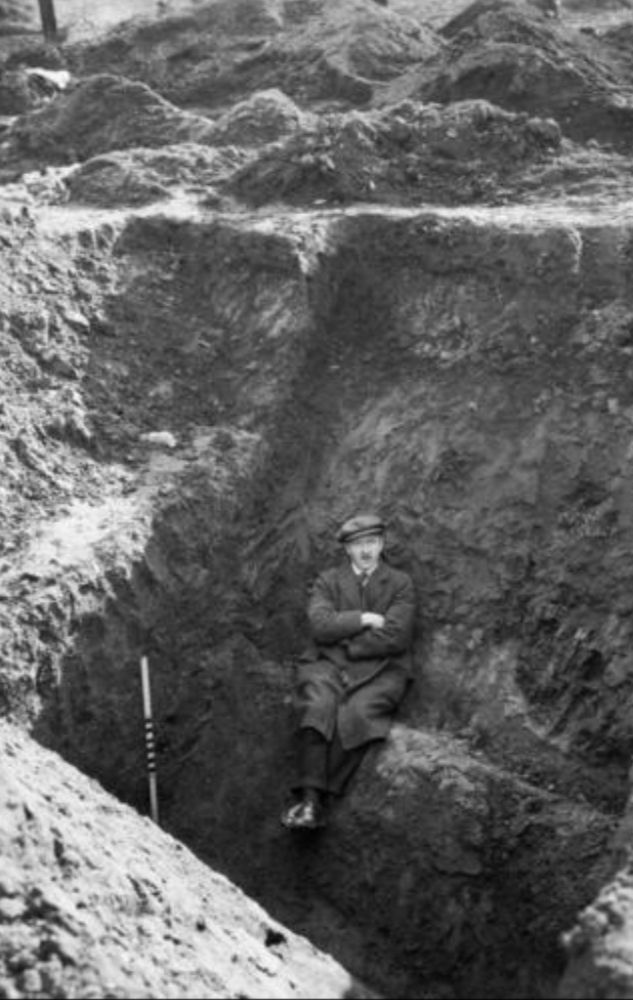 A black and white photo of a man in a suit and flat cap sitting on a ledge on the side of an enormous muddy ditch during excavations of Mumrills Roman fort.