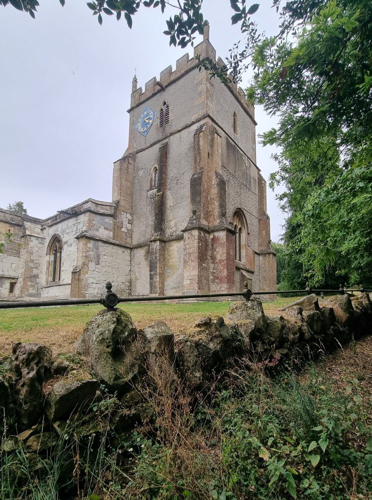 A line of large stones forming a sort of wall. Behind them is the church in Ashbury in Oxfordshire, which has a tall tower and a blue clock.