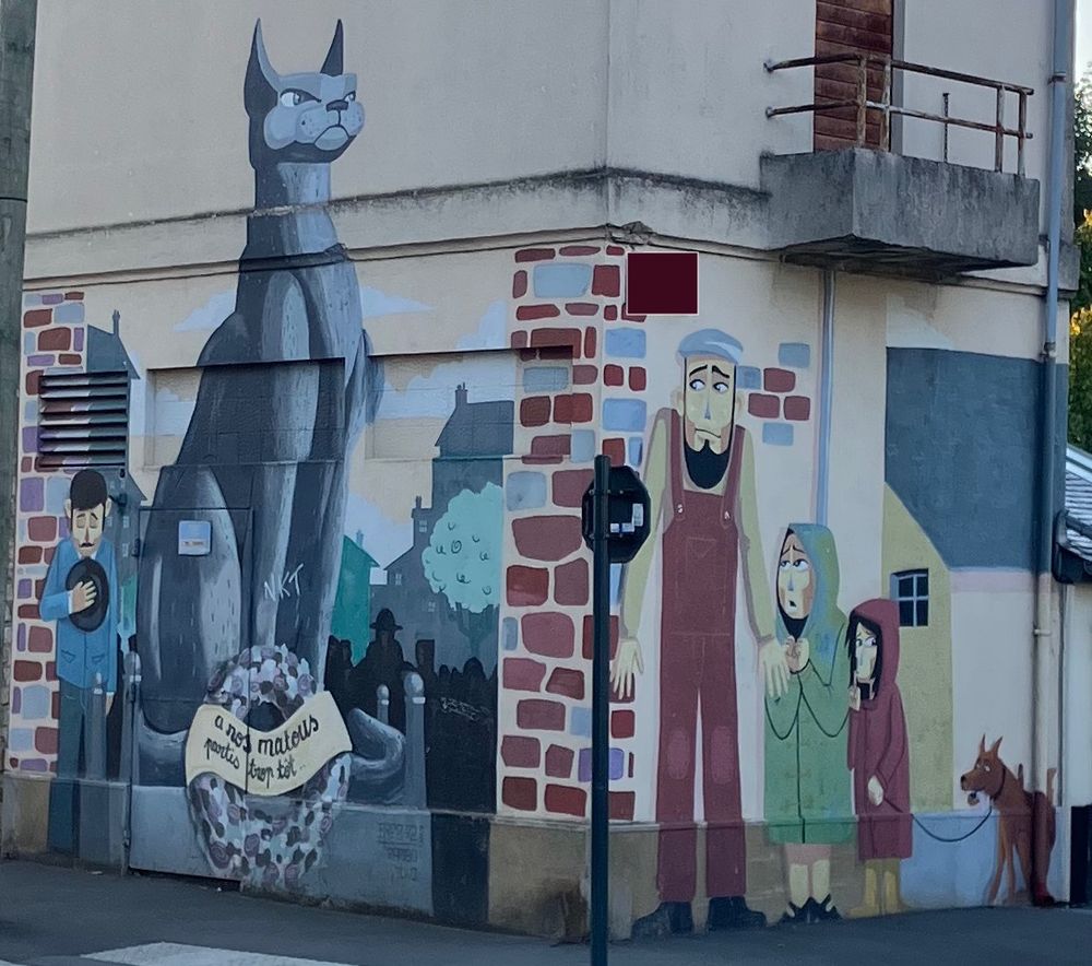 Une grande fresque à un coin de rue, d'un côté une statue de chat dépassant le première étage sur un mémorial. L'inscription dans la couronne mortuaire dit "à nos matous partis trop tôt". Sur l'autre côté, un homme en salopette apeuré avec deux enfants effrayés et un chien, un peu corniaud, en laisse.
