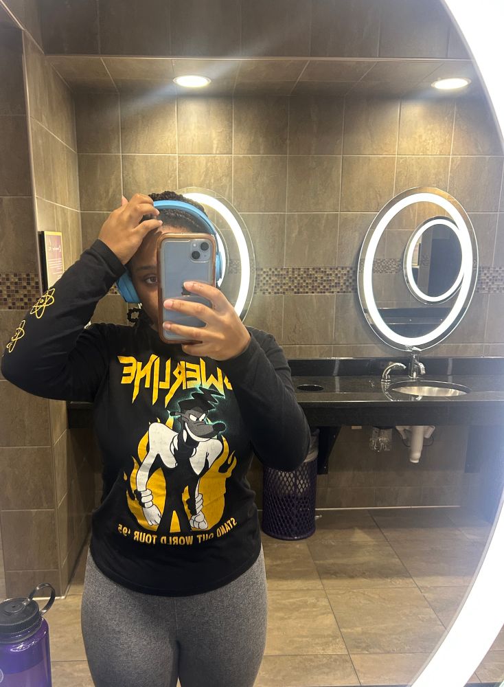 Woman wearing grey leggings, a long sleeved black tour shirt depicting power line from the first goofy movie, blue beats headphones, holding a blue iPhone aloft in her right hand in a gym bathroom. A purple water bottle rests on the counter in front of her. Her left hand is scratching at her hairline. Her phone obscures her face with the exception of one eye and eyebrow.