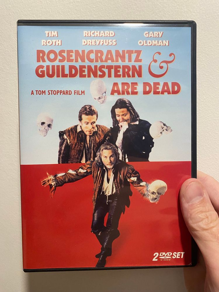 Cover of the DVD package for Rosencrantz and Giildenstern Are Dead. Foreground is Richard Dreyfuss doing a bow with a human skull in his outstretched left hand on a red background. Behind him, standing as if looking down at him from behind are Tim Roth and Gary Oldman with several Homan skulls floating around them.