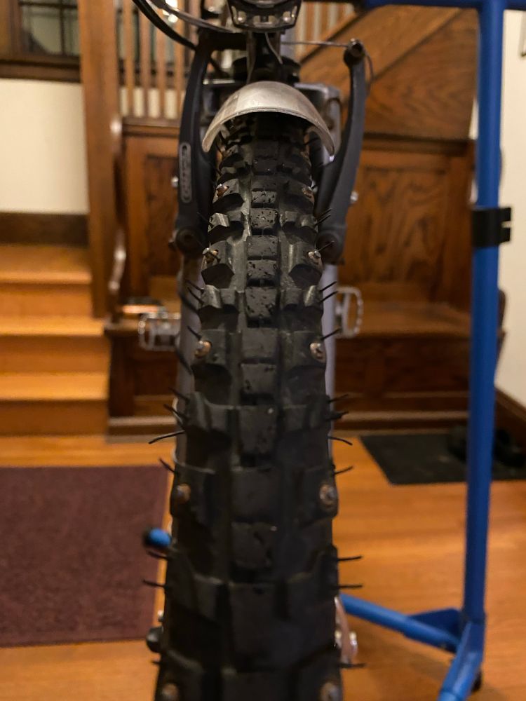 Closeup shot of a knobby bike tire with carbide-tipped metal studs on the two outer rows of knobs. The bike is on a blue Park Tools bike stand in the entryway to a residence.