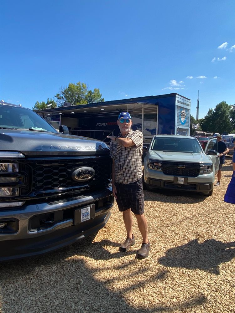 Man of 5´10 wearing shorts, plaid shirt and blue cycling cap standing next to the grill of a Ford F-something pickup truck with the hood coming up to his shoulder height.