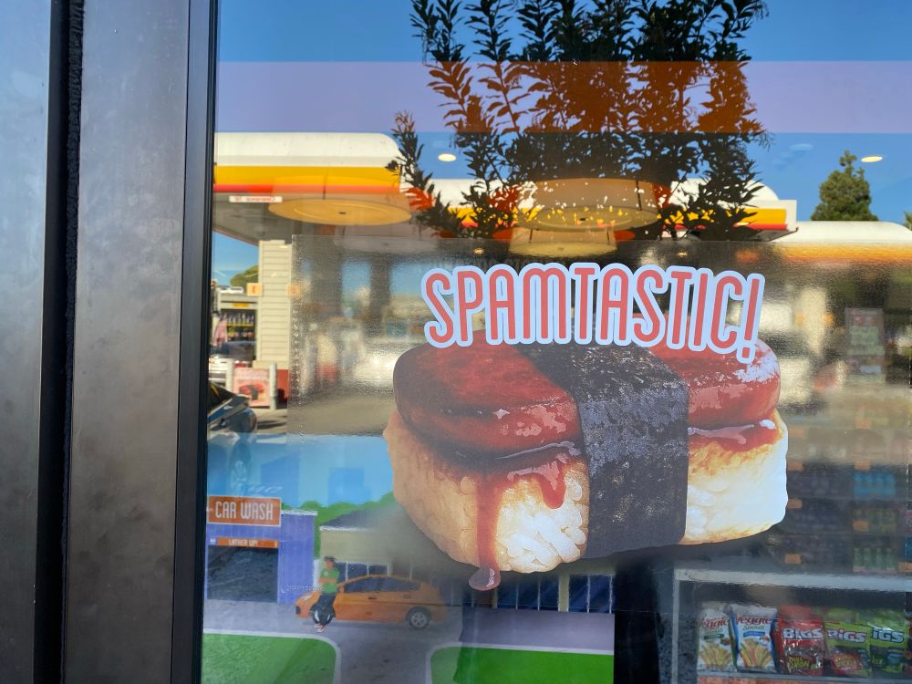 Advertisement sticker on the glass door of a service station of what looks like a piece of sushi with rice on the bottom and a piece of spam on top wrapped with a strip of seaweed. The word « Spamtastic » sits atop the erstwhile sushi.