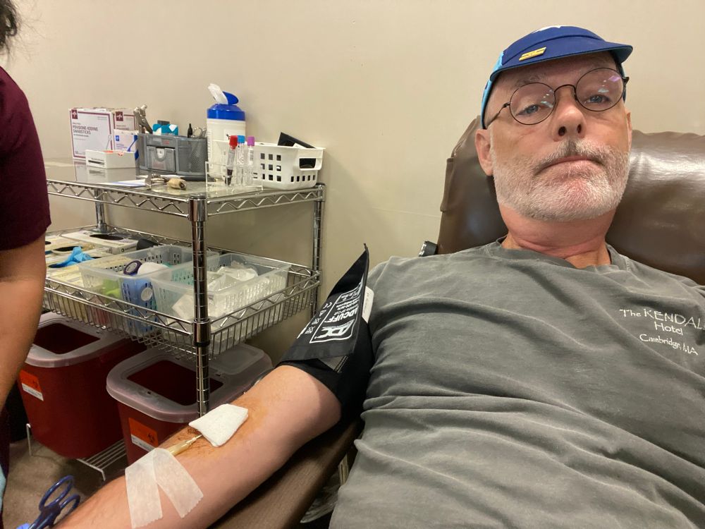 Grey-bearded man in a blue bike cap and grey shirt is reclining on a chaise with a blood donation rig hooked up to his right arm. A cart full of clinic supplies sits next to him.