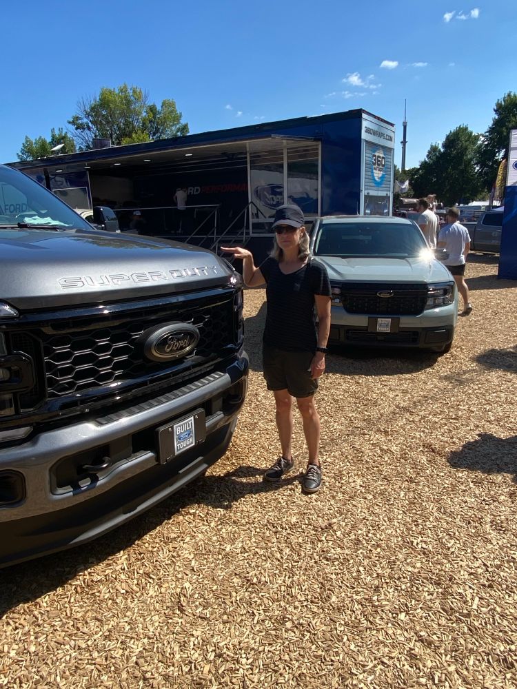 Woman of 5’3 wearing black shorts and shirt in black baseball cap standing in front of the grill of the same pickup truck, with the hood coming up to her chin. 