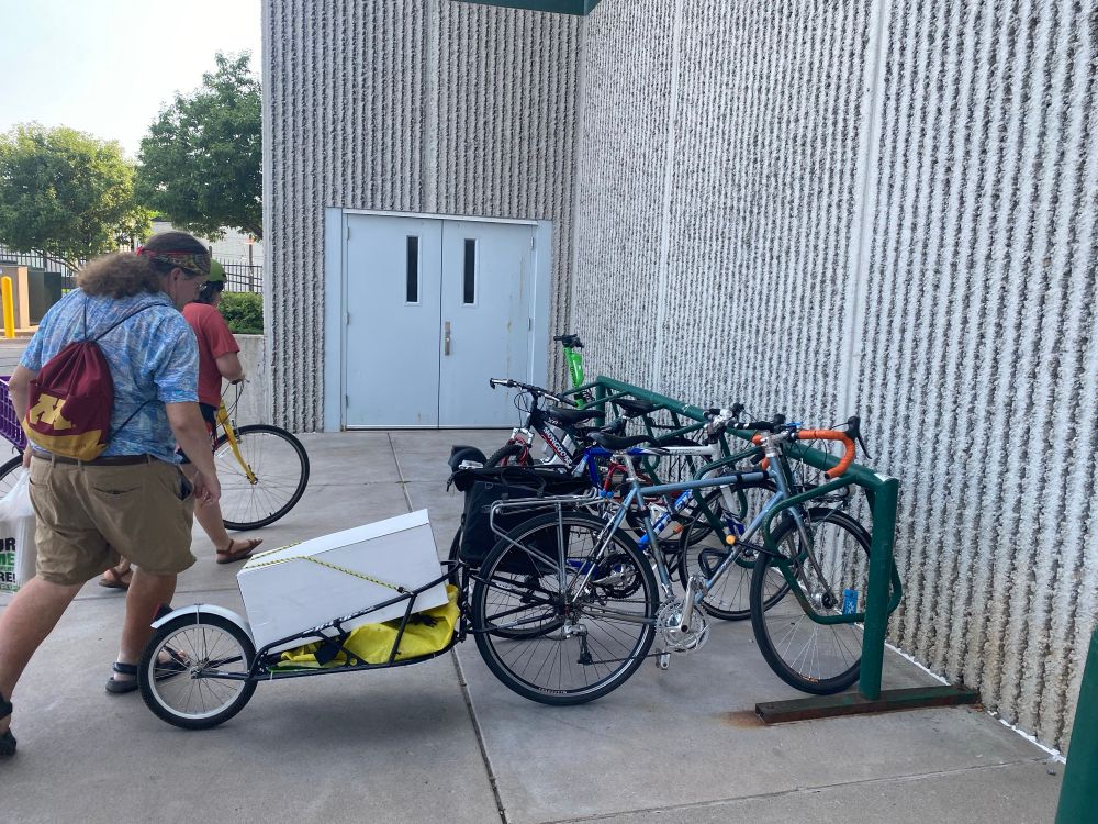 Full bikerack in front of a Menards store. Nearest bike has a B.O.B trailer carrying a white box that is roughly 12x12x24”. Behind this is one person walking towards the rack to take their bike and another person rolling their bike up to lock it. 