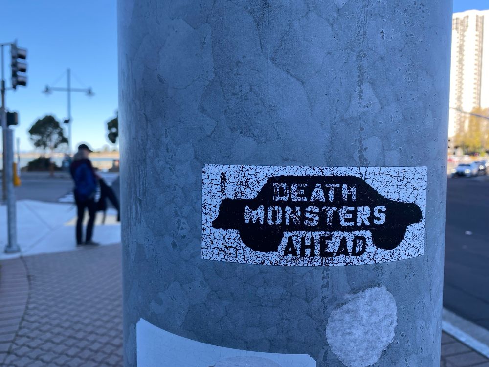 Sticker on a traffic light post with silhouette of a sedan style car and words « Death Monsters Ahead »