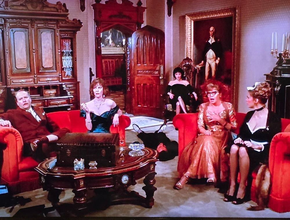 A picture of a scene from the film, Clue. Colonel Mustard and Ms. Scarlet are sitting on a red sofa while Mrs. Peacock and Yvette are sitting next to them on a red loveseat. 
Mrs. White in a seat in the background, with the corpse of Mr
Body lying on the ground in front of her.