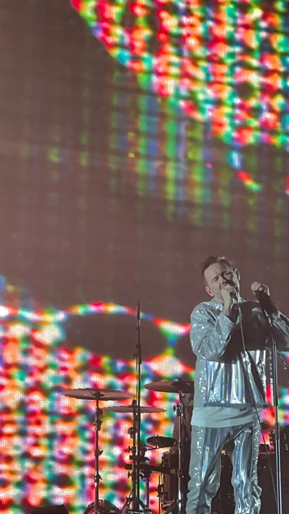 Joakim Berg i glittriga scenkläder står nere till höger i bild och sjunger med inlevelse mot en bakgrund av ett trumset och en stor glitchy backdrop/bildskärm.